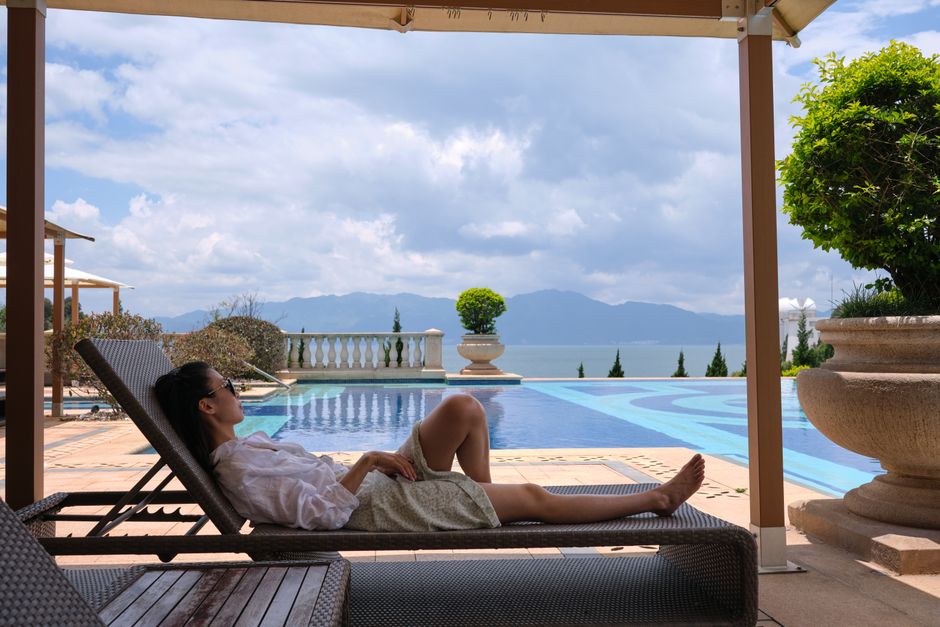 A woman in sunglasses relaxes on a sunlounger by a luxury poolside with a scenic view.